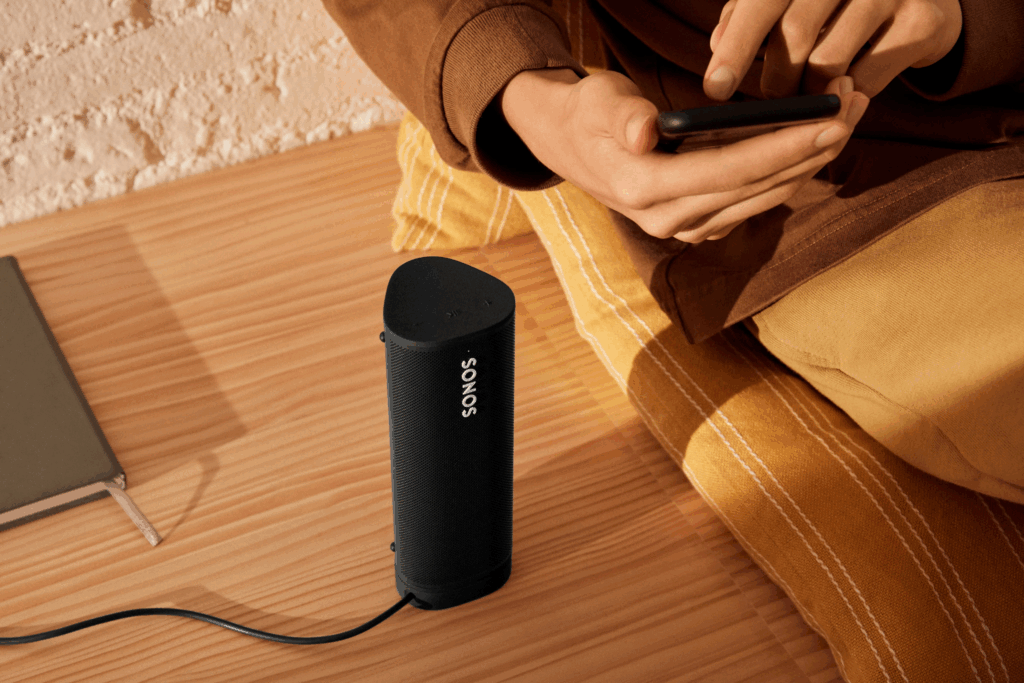 Close-up of a black, cylindrical Sonos speaker sitting on a light wooden floor, with a man wearing a brown and yellow outfit seated next to it, scrolling on his smartphone. The lighting has a warm, yellow-orange glow against a textured white/black brick wall, emphasizing the speaker as the ultimate portable music gift for the tech-savvy man.