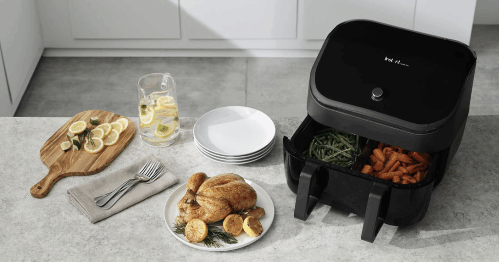A black Instant Vortex dual-basket air fryer with the drawers pulled open to show green beans and carrots. A roasted chicken sits on a white plate to the left of the unit.