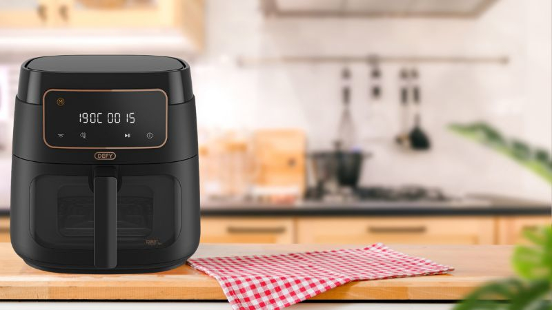 A black Defy air fryer with a transparent viewing window and rose-gold trim, sitting on a wooden countertop next to a red and white checkered cloth.