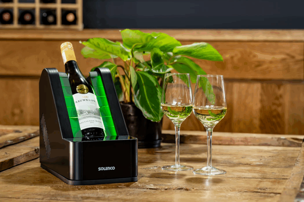 Solenco Electric Wine Chiller Lifestyle: A black, modern Solenco rapid wine chiller containing a bottle of white wine, illuminated by a bright, vibrant green light, sits on a rustic wooden table. Two stemmed glasses filled with golden-yellow wine stand beside it, creating a sophisticated and luxurious tableau. This is the perfect practical gift for the man who appreciates perfectly chilled drinks.